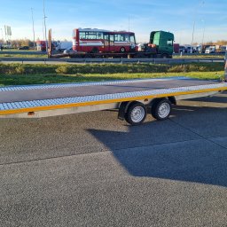 Rafał Rebicki Firma Handlowo Szkoleniowa err - Pusta, dwuosiowa laweta samochodowa z najazdami, zaparkowana na parkingu. W tle autobus na lawecie i inne pojazdy ciężarowe. Słoneczny dzień.