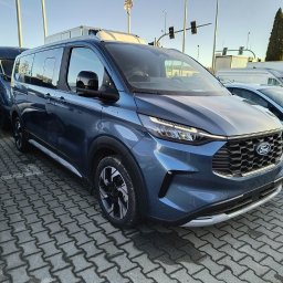 Rafał Rebicki Firma Handlowo Szkoleniowa err - Niebieski Ford Transit Custom w Somoninie, zaparkowany na kostce brukowej. Widoczne inne pojazdy w tle, w tym busy. Ujęcie z przodu, lekko z boku.