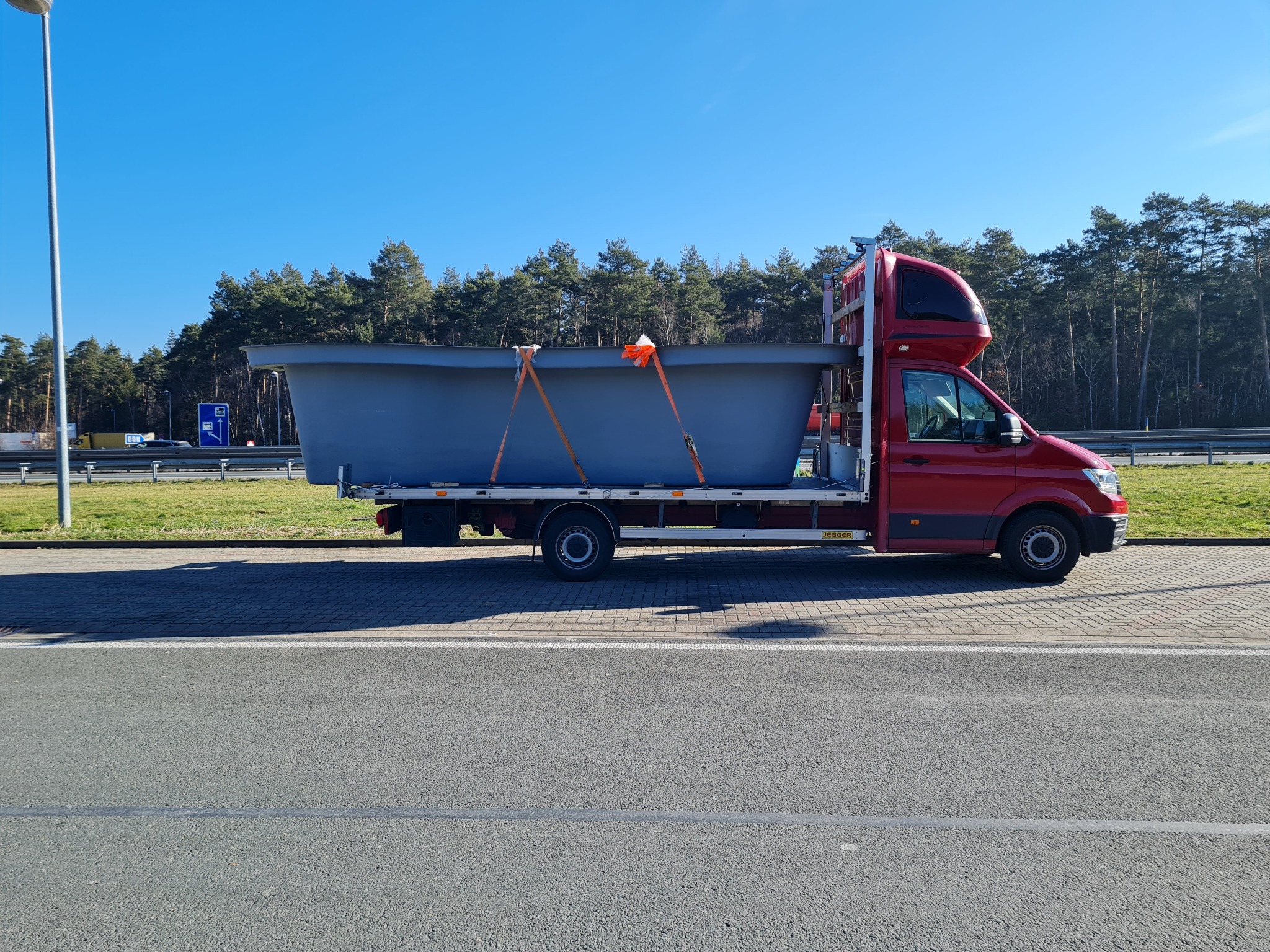 Czerwony bus z kabiną sypialną przewozi szary, duży zbiornik zabezpieczony pasami transportowymi. Widok z boku, w tle drzewa i błękitne niebo.