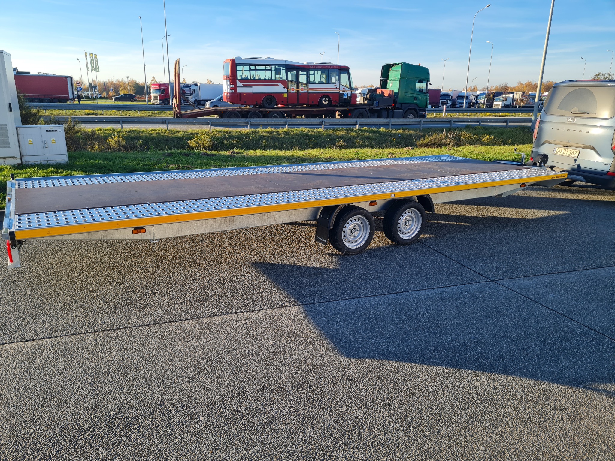 Pusta, dwuosiowa laweta samochodowa z najazdami, zaparkowana na parkingu. W tle autobus na lawecie i inne pojazdy ciężarowe. Słoneczny dzień.