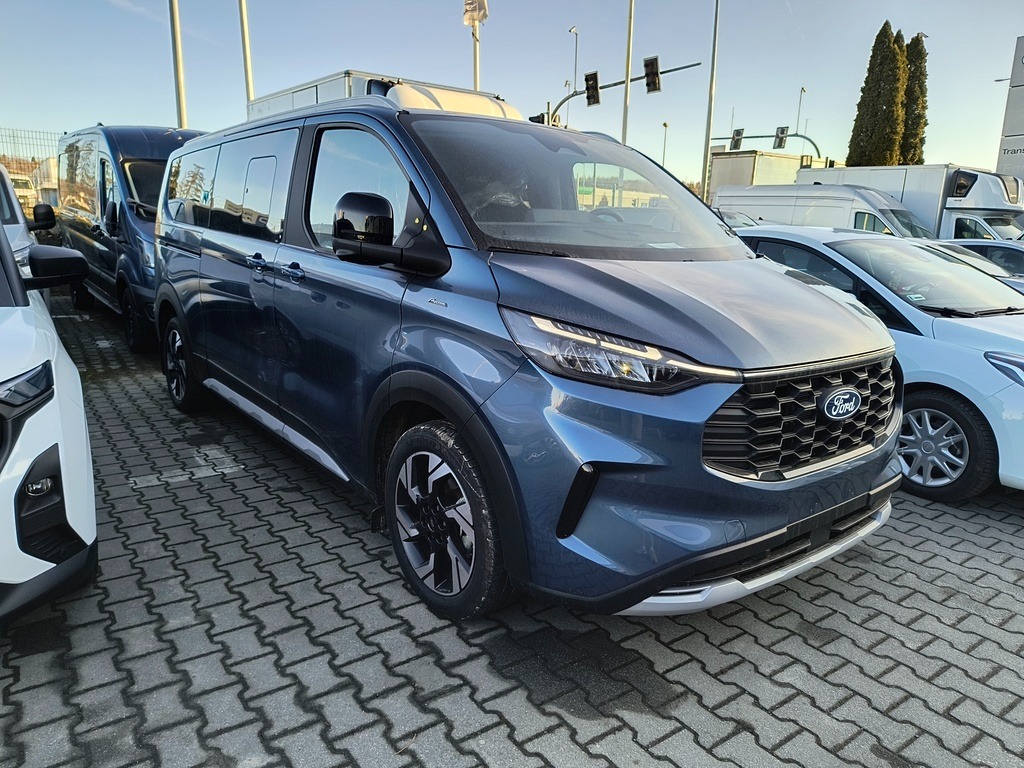 Niebieski Ford Transit Custom w Somoninie, zaparkowany na kostce brukowej. Widoczne inne pojazdy w tle, w tym busy. Ujęcie z przodu, lekko z boku.