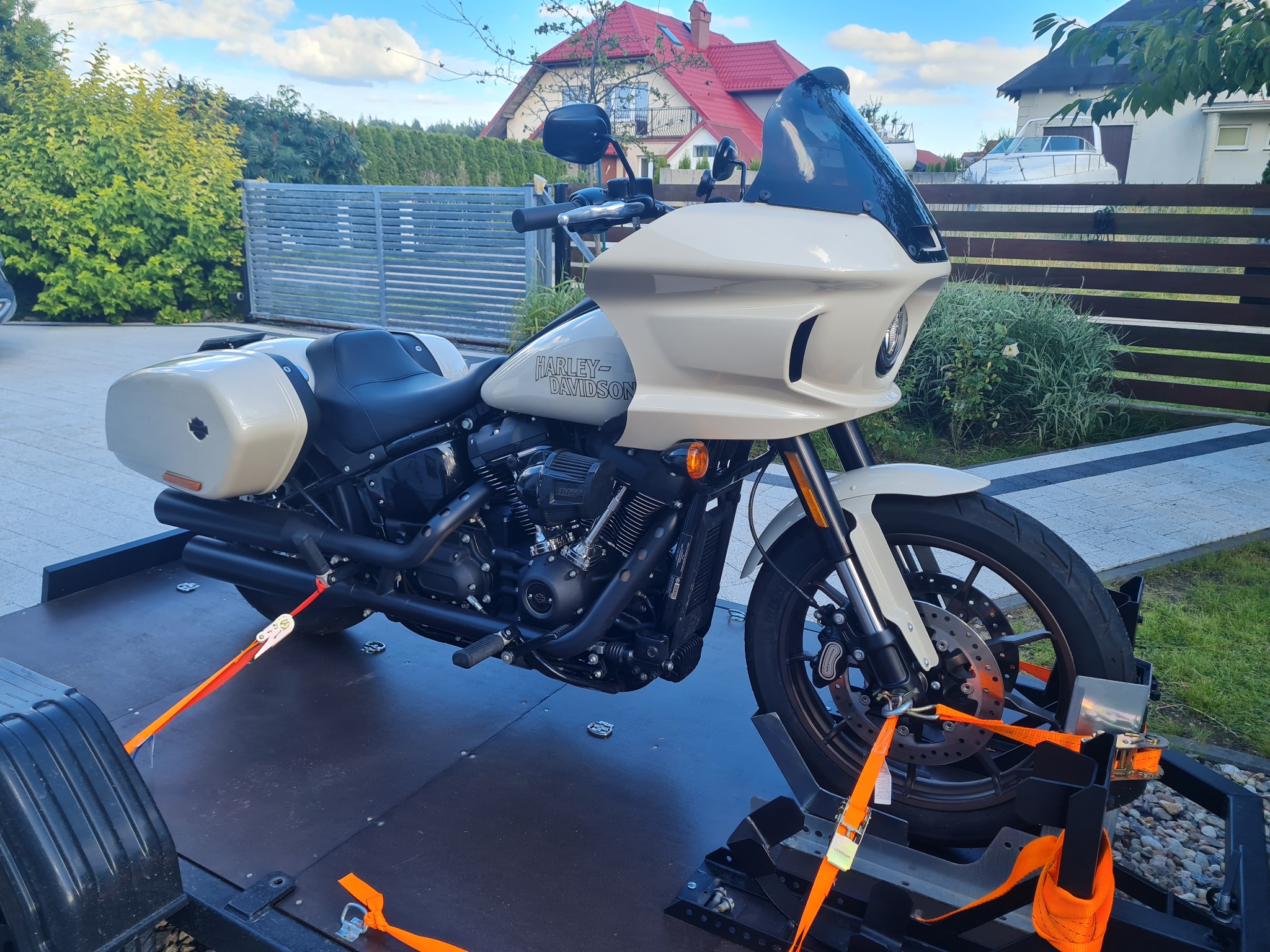 Kremowy motocykl Harley-Davidson na czarnej lawecie, zabezpieczony pomarańczowymi pasami transportowymi. W tle dom z czerwoną dachówką i zielony ogród.