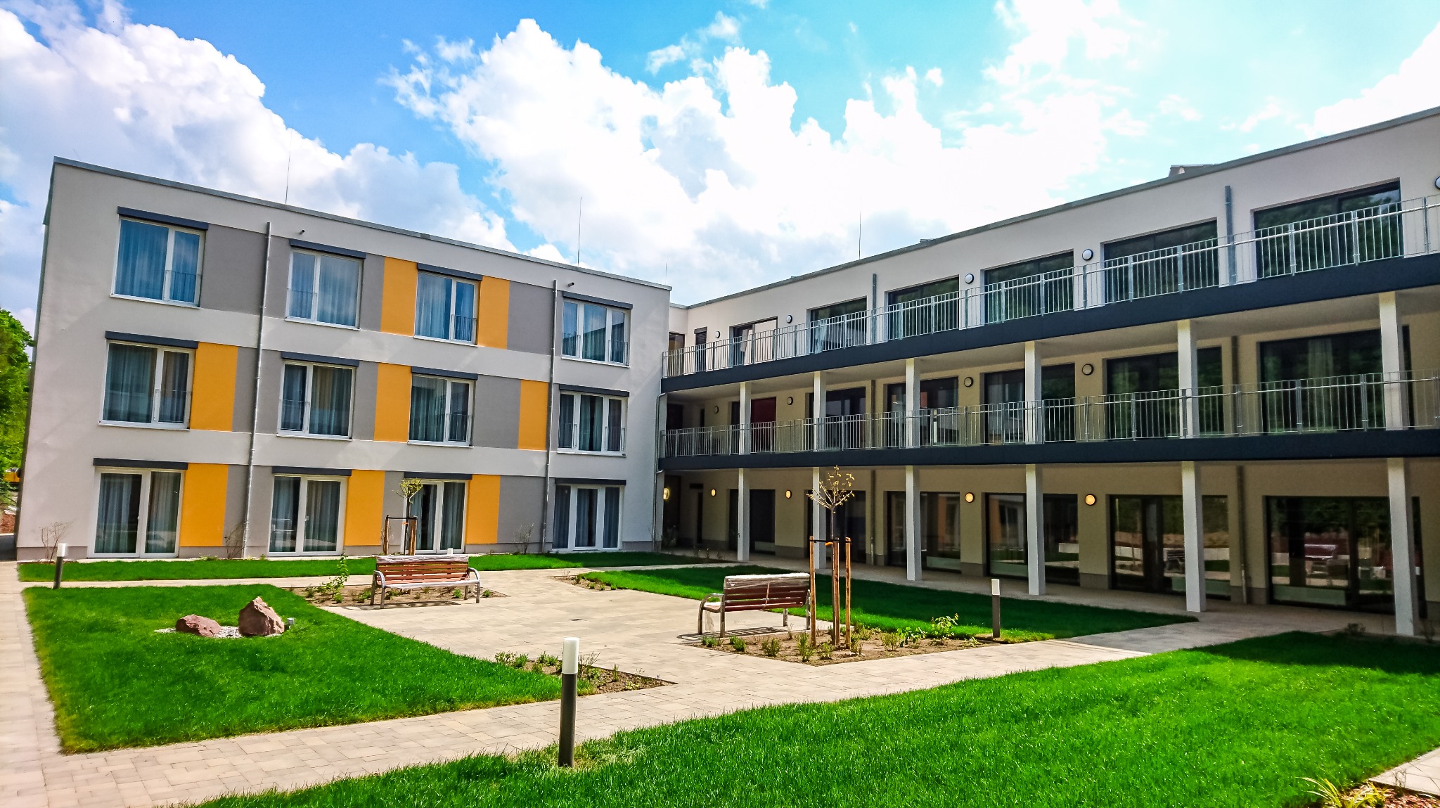 Dziedziniec nowoczesnego budynku wielorodzinnego z zielonym trawnikiem, ścieżką z kostki i ławkami. Budynek w Hainichen z balkonami i elementami w kolorze szarym i żółtym.