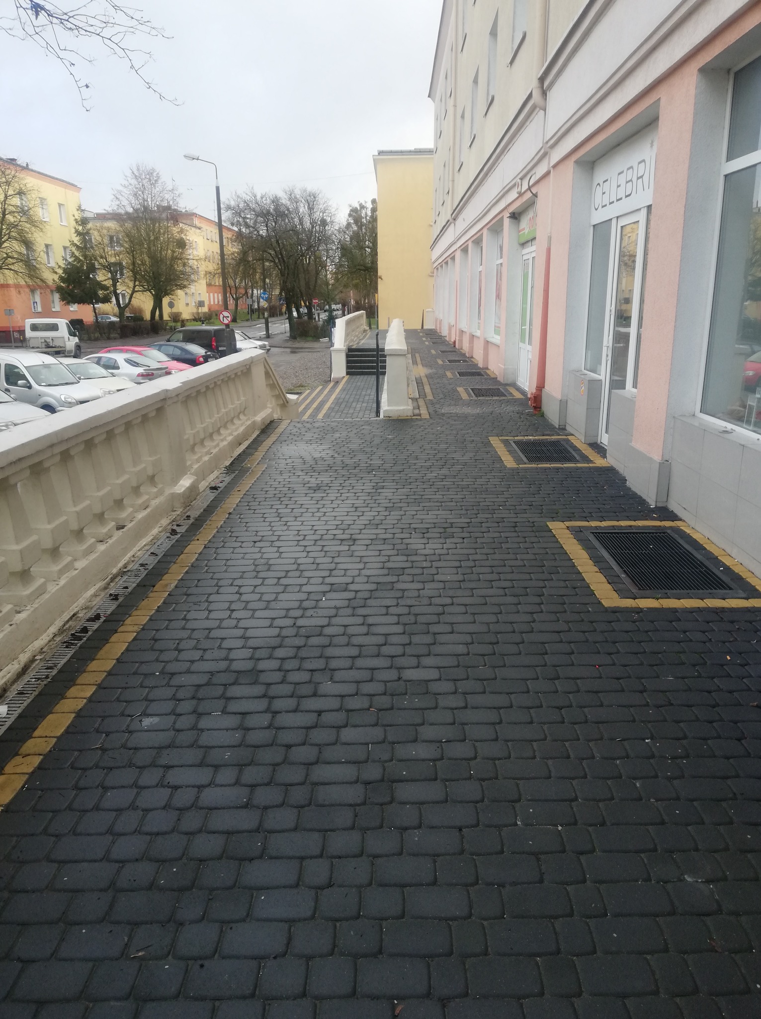 Czarna kostka brukowa z żółtymi akcentami, ułożona wzdłuż budynku z oknami i balustradą. Widoczne studzienki kanalizacyjne z żółtymi obramowaniami.