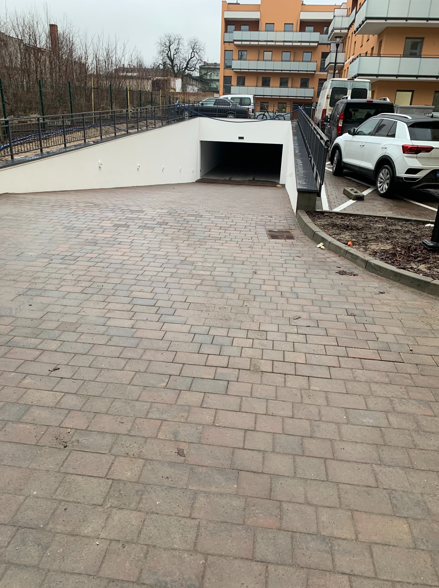 Ułożona kostka brukowa prowadząca do wjazdu pod budynek. Widoczne zaparkowane samochody i fragment budynku mieszkalnego w tle. Kostka w odcieniach szarości i brązu.