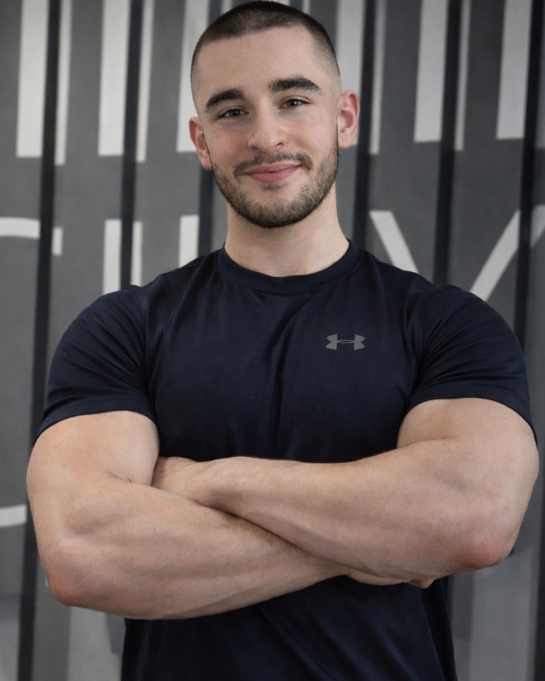 Uśmiechnięty mężczyzna w czarnej koszulce sportowej z założonymi rękami na tle szaro-białej ściany. Krótkie włosy, zadbana broda. Dobre oświetlenie.