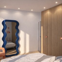 DRZEWIECKA DESIGN - Sypialnia z dużą szafą w odcieniach drewna, falistym lustrem w niebieskiej ramie i pomarańczowym fotelem. Minimalistyczny design, naturalne światło.