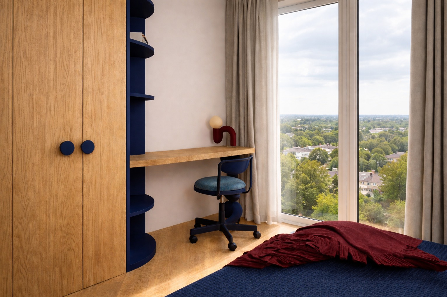 Pokój z biurkiem i szafą w zabudowie, utrzymany w tonacji drewna i granatu. Za oknem widok na zieleń. Nietypowa lampka na biurku dodaje charakteru.