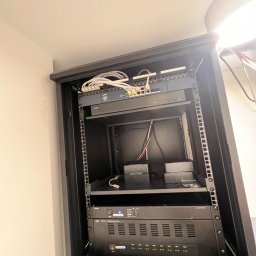 Doorox Dominik Steckert - Zamknięta szafa rack z zainstalowanymi urządzeniami sieciowymi i wzmacniaczami audio, widoczne kable i diody sygnalizacyjne. Sprzęt w kolorze czarnym.