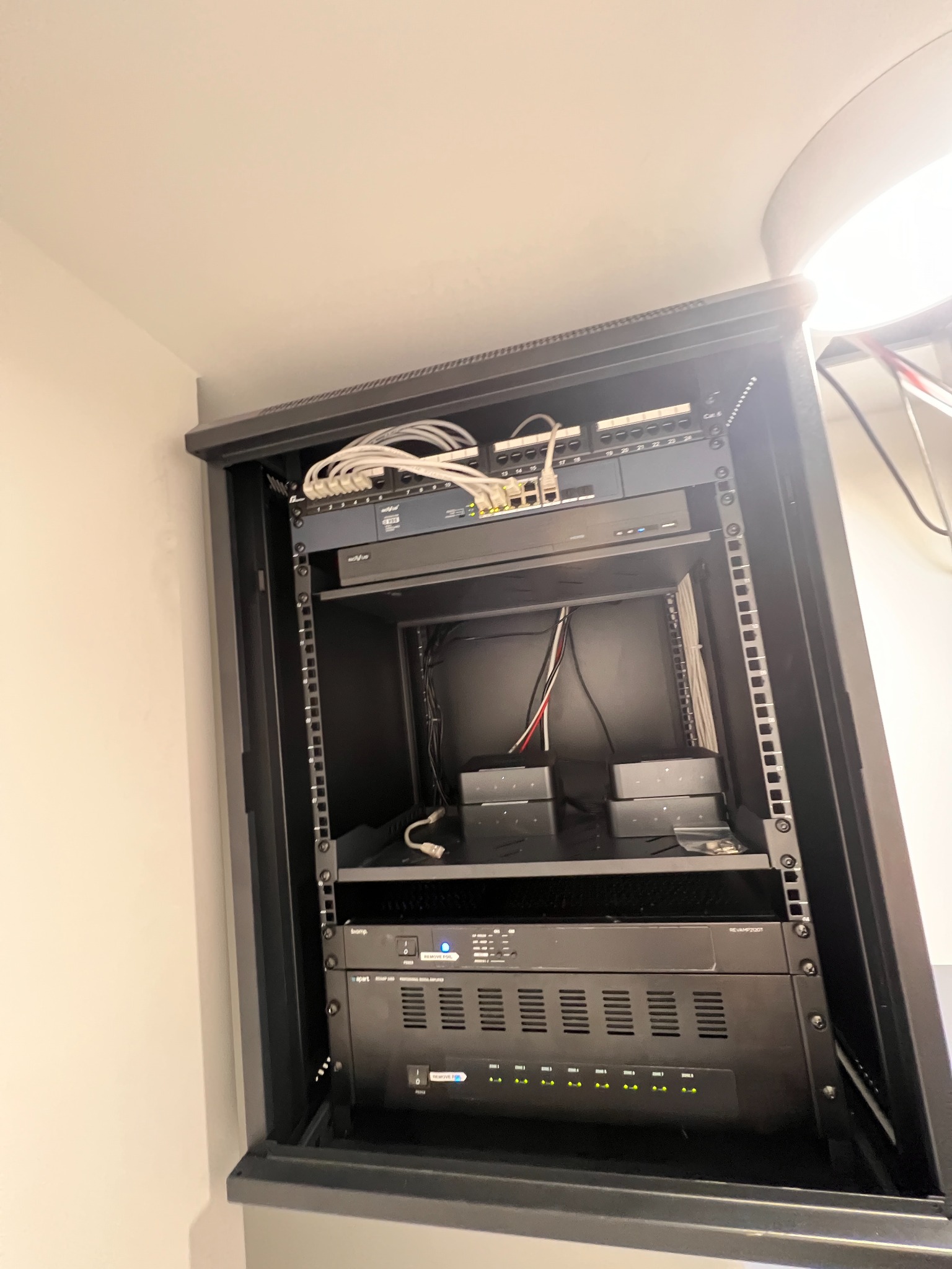 Zamknięta szafa rack z zainstalowanymi urządzeniami sieciowymi i wzmacniaczami audio, widoczne kable i diody sygnalizacyjne. Sprzęt w kolorze czarnym.