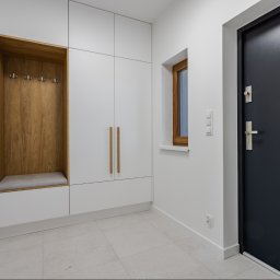 Chata na wymiar Sp. z o.o. - Minimalistyczna zabudowa przedpokoju z siedziskiem i wieszakami, białe szafki z drewnianymi uchwytami, szara podłoga, czarne drzwi wejściowe z przeszkleniem.