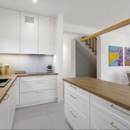 Chata na wymiar Sp. z o.o. - Kremowa kuchnia z drewnianym blatem i wyspą. Nowoczesne fronty, minimalistyczne uchwyty, jasne wnętrze z elementami dekoracyjnymi i schodami w tle.