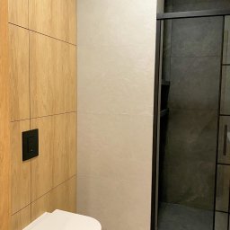 Chata na wymiar Sp. z o.o. - Nowoczesna zabudowa WC na wymiar: drewnopodobne szafki, podwieszana toaleta z czarnym przyciskiem i kabina prysznicowa z ciemnymi płytkami.