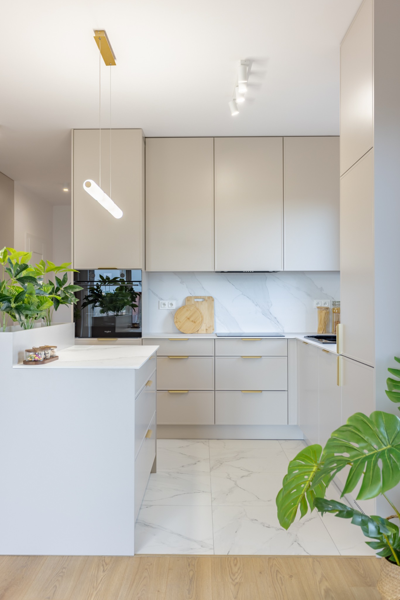 Jasna kuchnia w odcieniach kaszmiru z marmurowym blatem i złotymi uchwytami, oświetlona nowoczesną lampą. Rośliny doniczkowe dodają wnętrzu świeżości.
