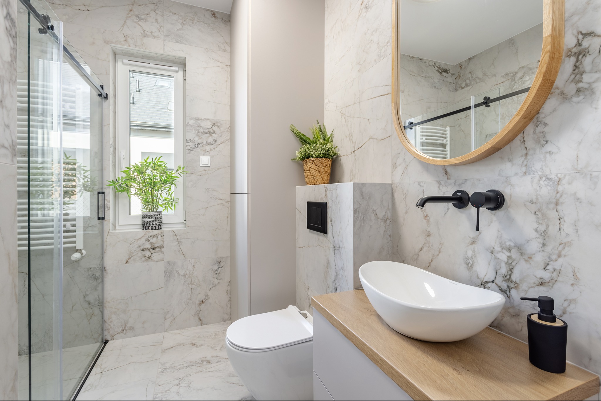 Elegancka łazienka z marmurowymi płytkami, owalnym lustrem w drewnianej ramie, białą umywalką nablatową i czarną armaturą. Roślina w oknie dodaje świeżości.