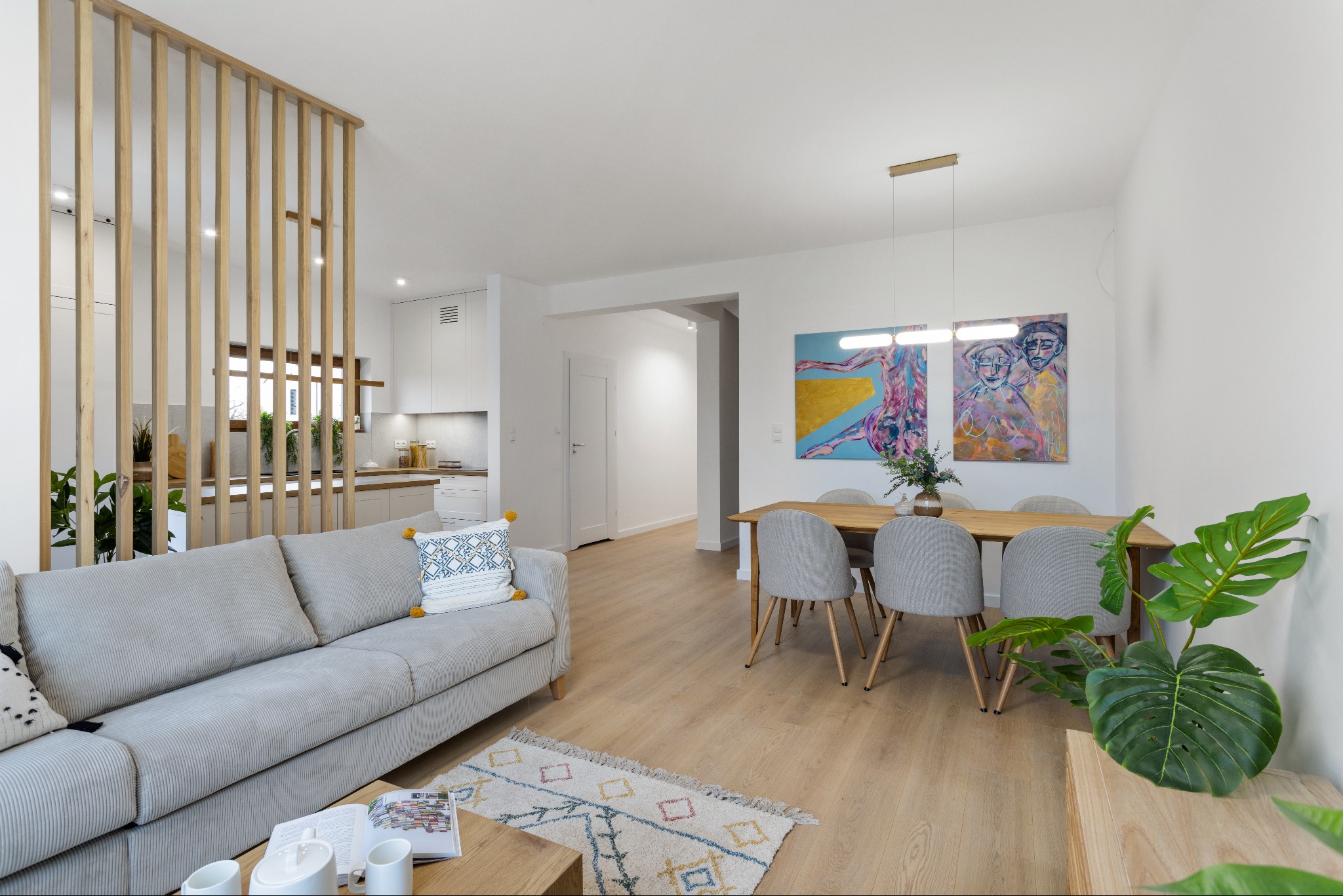 Jasne wnętrze: salon z szarą sofą, drewnianym stołem i lamelami oddzielającymi kuchnię. Nowoczesny design, obrazy na ścianie, roślina w rogu.