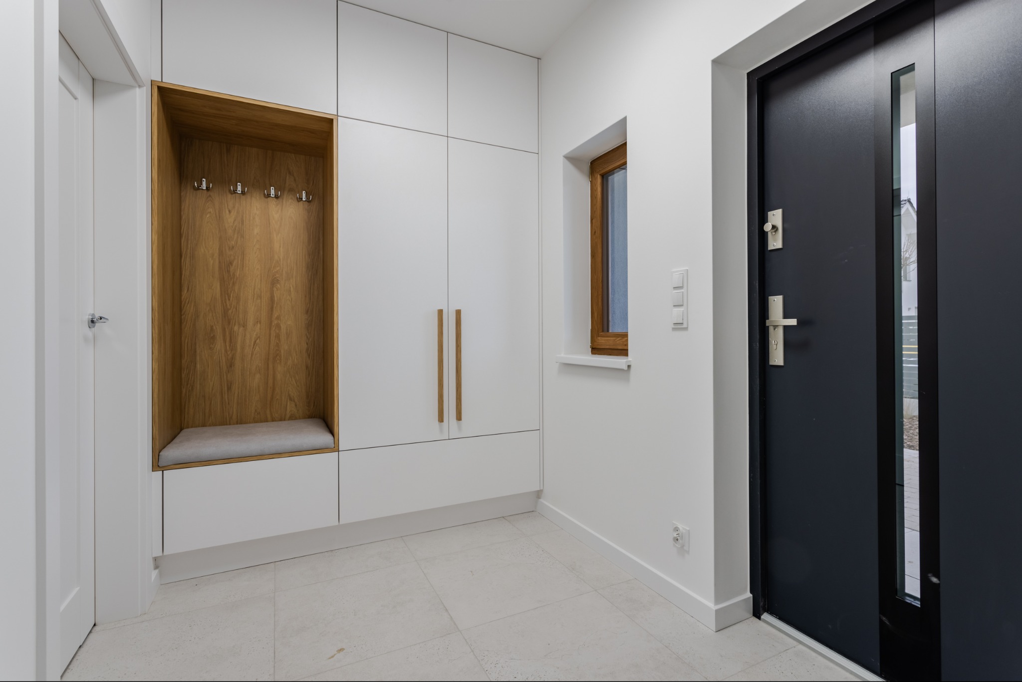 Minimalistyczna zabudowa przedpokoju z siedziskiem i wieszakami, białe szafki z drewnianymi uchwytami, szara podłoga, czarne drzwi wejściowe z przeszkleniem.