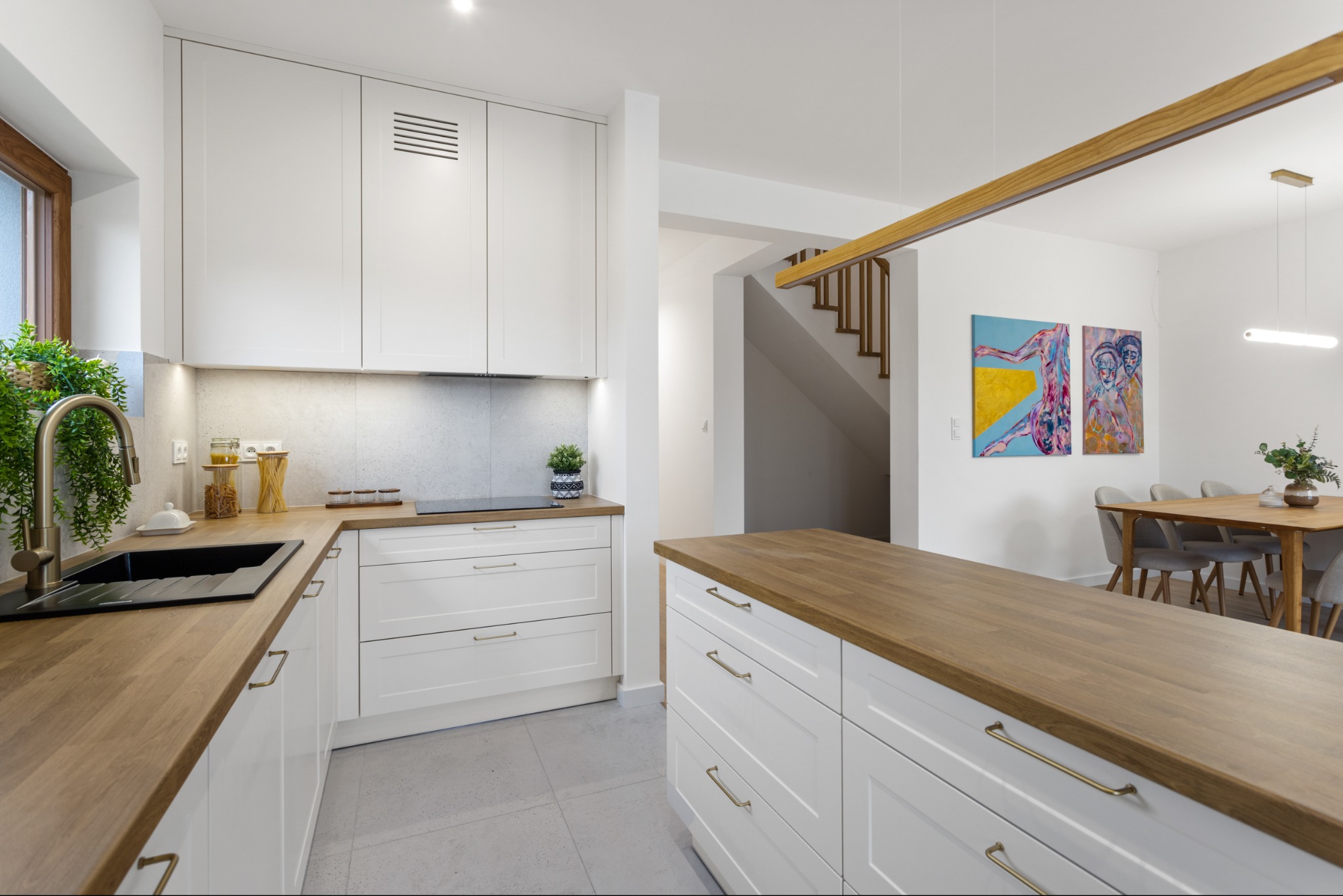 Kremowa kuchnia z drewnianym blatem i wyspą. Nowoczesne fronty, minimalistyczne uchwyty, jasne wnętrze z elementami dekoracyjnymi i schodami w tle.