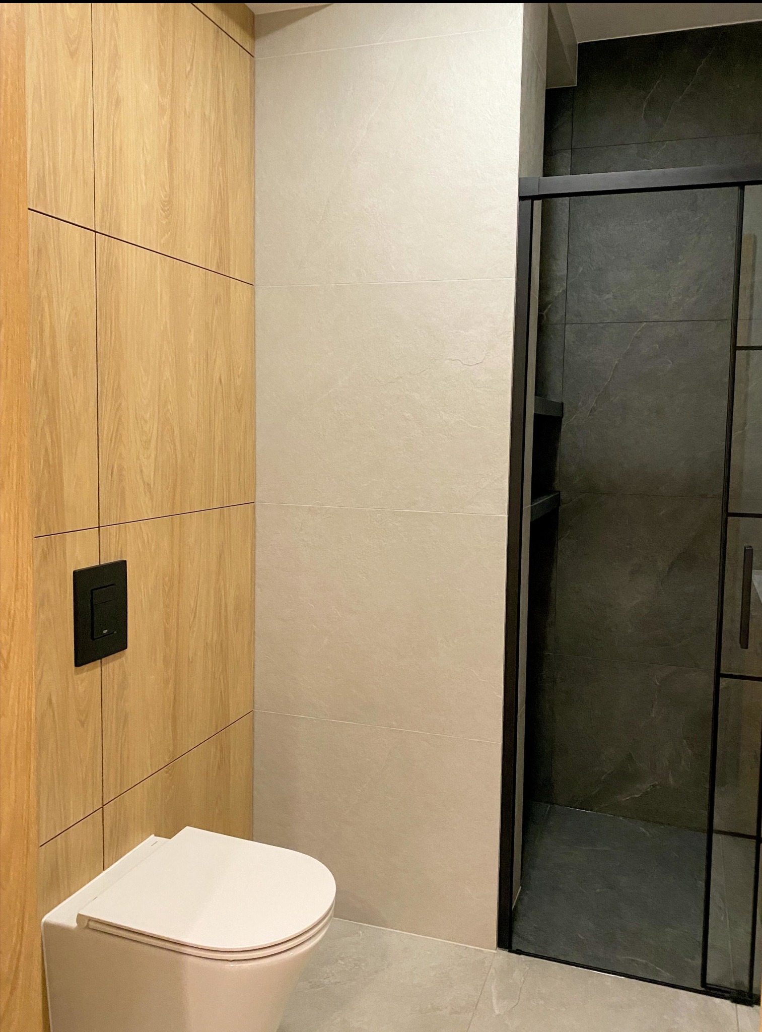 Nowoczesna zabudowa WC na wymiar: drewnopodobne szafki, podwieszana toaleta z czarnym przyciskiem i kabina prysznicowa z ciemnymi płytkami.