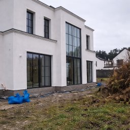 Fillpro Ivan Filiarovskyi - Biała elewacja domu z dużymi oknami w Nadarzynie. Widoczne detale architektoniczne i przygotowany teren wokół budynku. Nowoczesny styl.
