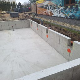 M&P Sp. zo.o. - Żelbetowy fundament basenu w ziemi, z widocznymi otworami na instalacje. W tle budynek z muralem i pracownicy budowlani. Surowy beton, świeżo wylany.