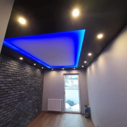 Czad Bud Kinga Sadowa - Pokój po remoncie: czarny sufit z podświetleniem LED, ściana z cegły, szare ściany, drewniana podłoga. Widoczne okno i grzejnik. Nowoczesny design.