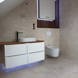 Czad Bud Kinga Sadowa - Nowoczesna łazienka na poddaszu z beżowymi płytkami, podświetlaną szafką pod umywalką z drewnianym blatem i wiszącą toaletą. Minimalistyczny design z akcentami drewna.