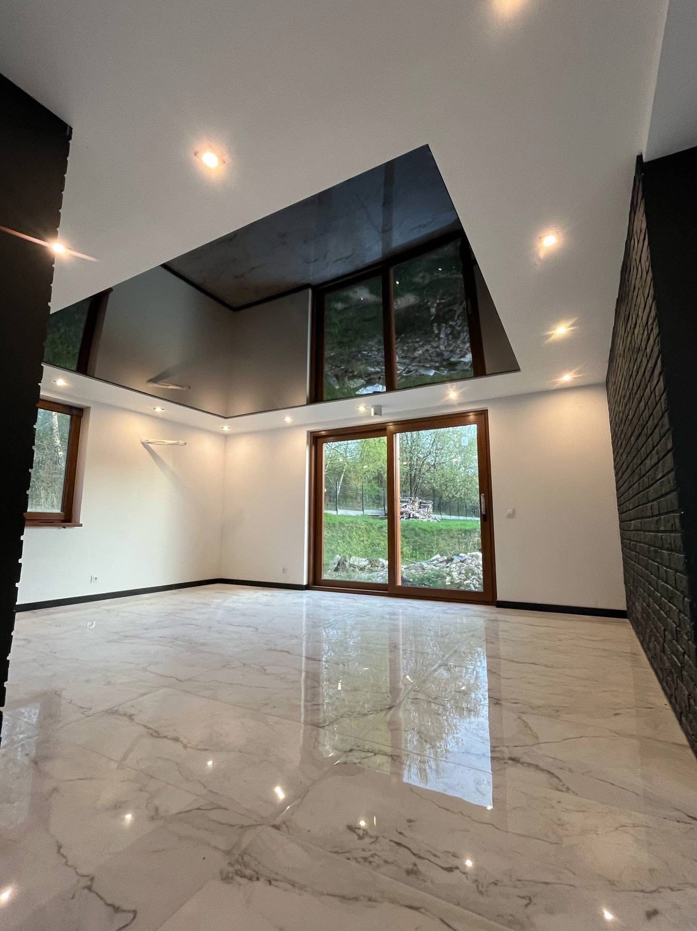 Nowoczesne wnętrze z marmurową podłogą i lustrzanym sufitem. Widoczne okna z widokiem na zieleń i czarna ściana z cegły. Oświetlenie punktowe podkreśla elegancję.