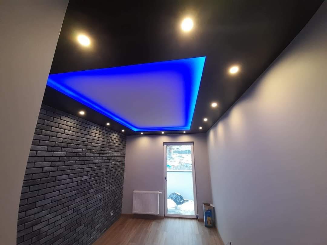 Pokój po remoncie: czarny sufit z podświetleniem LED, ściana z cegły, szare ściany, drewniana podłoga. Widoczne okno i grzejnik. Nowoczesny design.