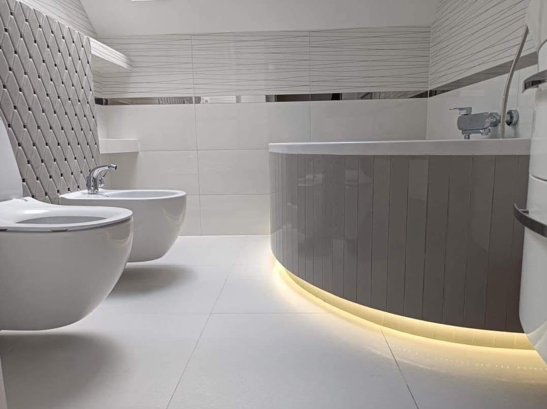 Nowoczesna łazienka w bieli i szarości z podświetlaną wanną, umywalką i bidetem. Geometryczne płytki na ścianie dodają unikalnego charakteru.