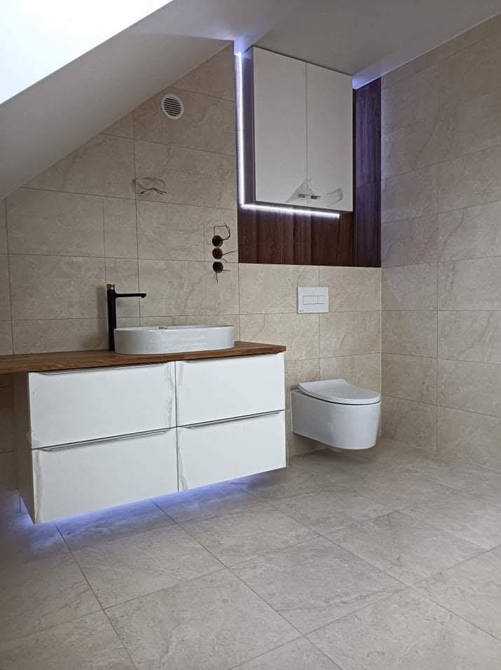 Nowoczesna łazienka na poddaszu z beżowymi płytkami, podświetlaną szafką pod umywalką z drewnianym blatem i wiszącą toaletą. Minimalistyczny design z akcentami drewna.