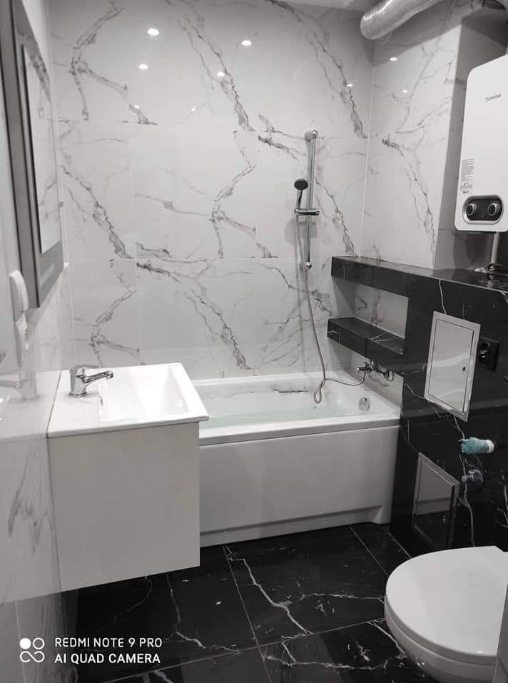 Wykończona łazienka z wanną, umywalką i toaletą. Białe marmurowe płytki na ścianach kontrastują z czarną marmurową podłogą. Nowoczesny design, oświetlenie punktowe.