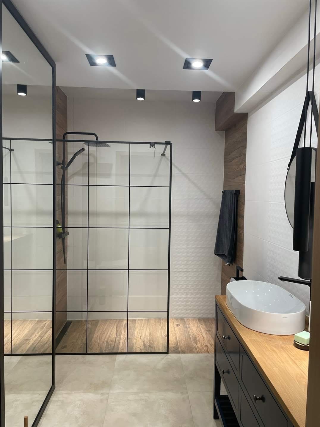Nowoczesna łazienka z kabiną prysznicową typu walk-in z czarnymi profilami, umywalką nablatową i drewnianymi akcentami. Minimalistyczny design, oświetlenie punktowe.