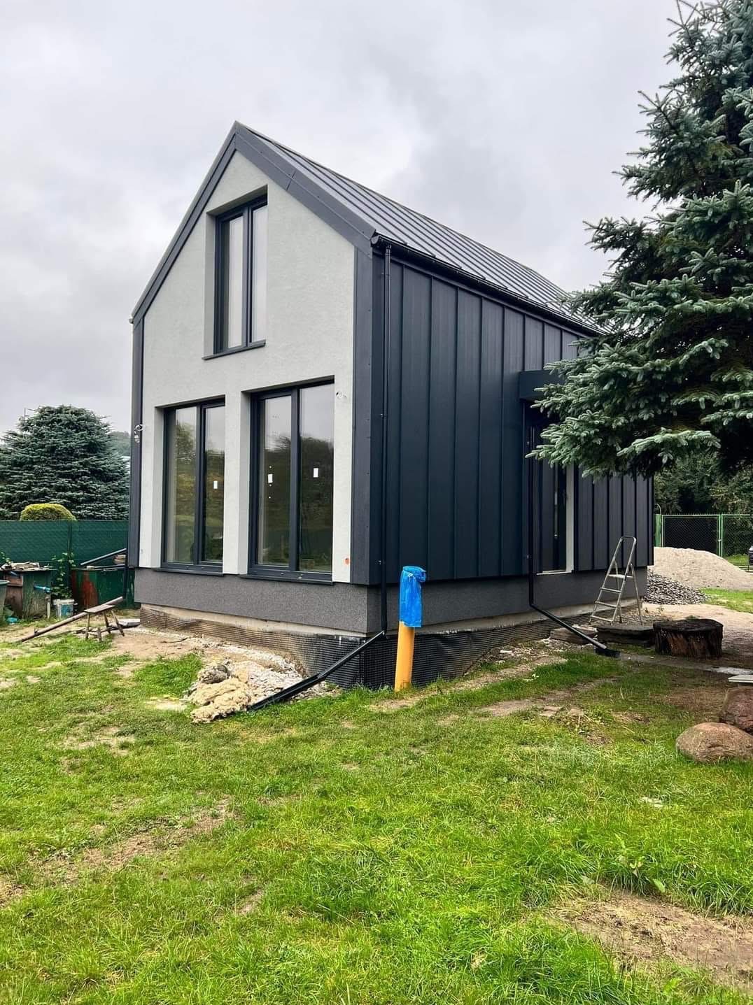 Nowoczesny dom z ciemną, pionową elewacją i jasnym tynkiem, widoczne okna z czarnymi ramami. W tle zieleń i pochmurne niebo. Na trawniku widoczne ślady prac.