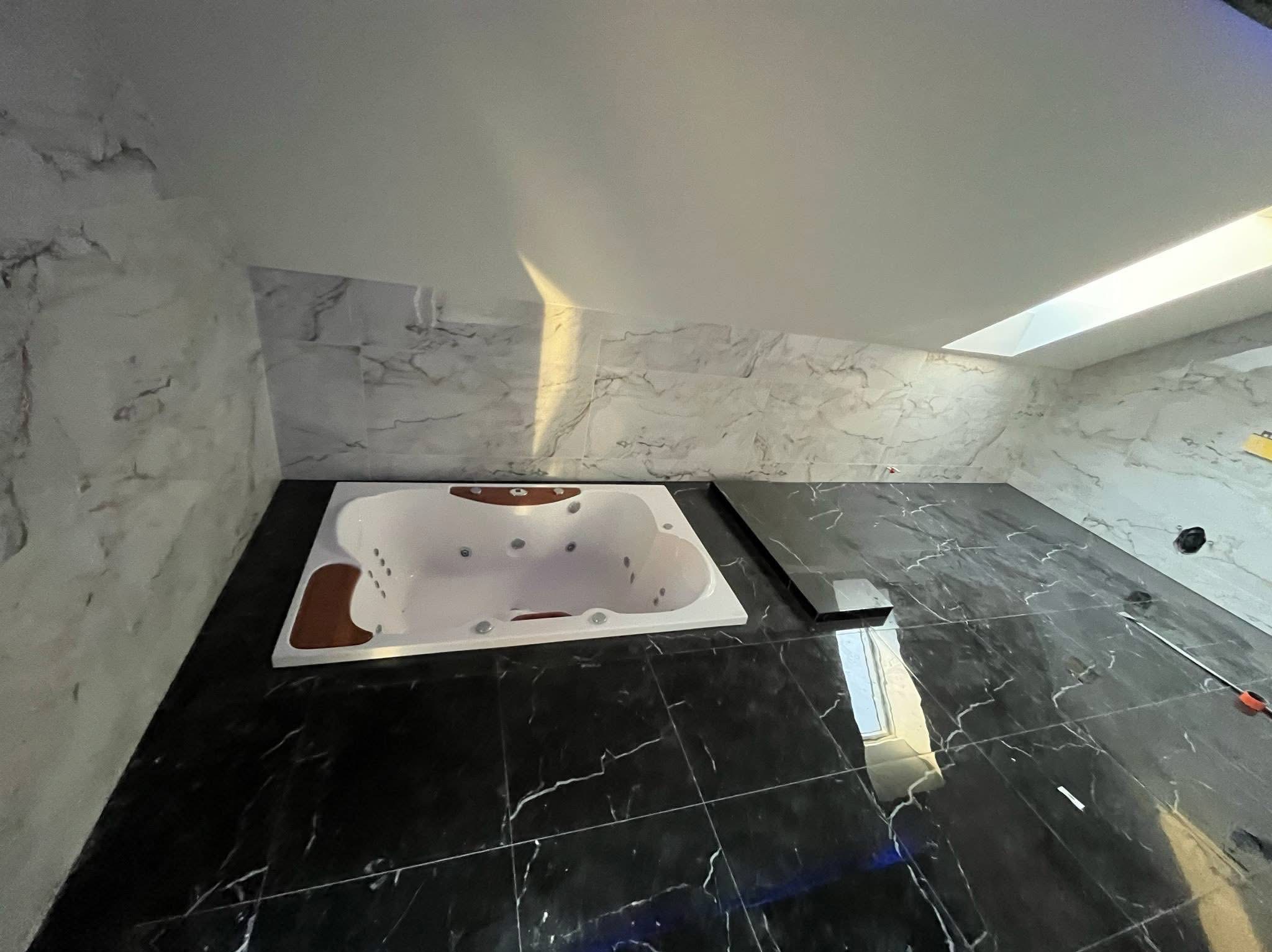 Nowoczesna łazienka z czarną marmurową podłogą i ścianami w odcieniach szarości. Biała wanna z hydromasażem wkomponowana w podłogę. Ukośny sufit z oknem.