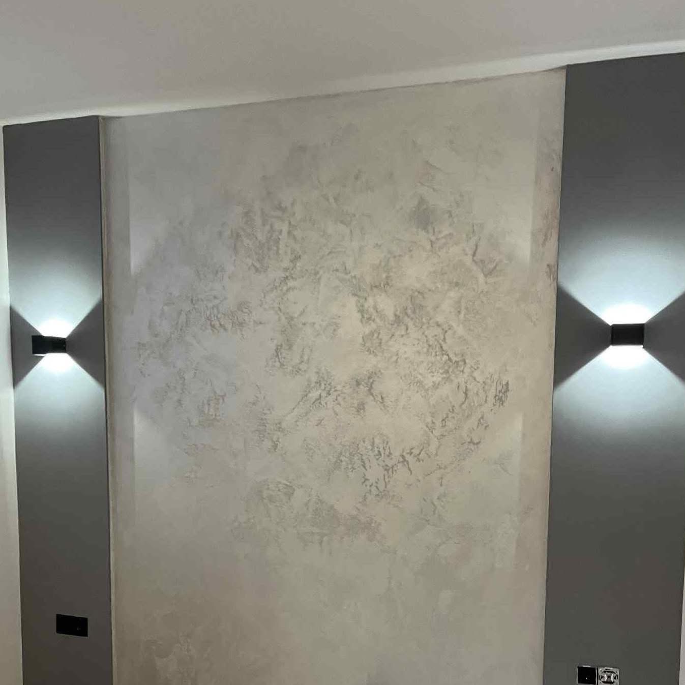 Dekoracyjna ściana z efektem stiuku w odcieniach szarości, oświetlona dwoma nowoczesnymi kinkietami. Widoczne gniazdka elektryczne. Subtelne przejścia tonalne.