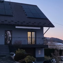 Audyty energetyczne. House Energy. Katarzyna Fejkiel - Szary dom jednorodzinny z panelami słonecznymi na dachu, numer 110, balkon z kutą balustradą, w tle góry i zachodzące słońce.