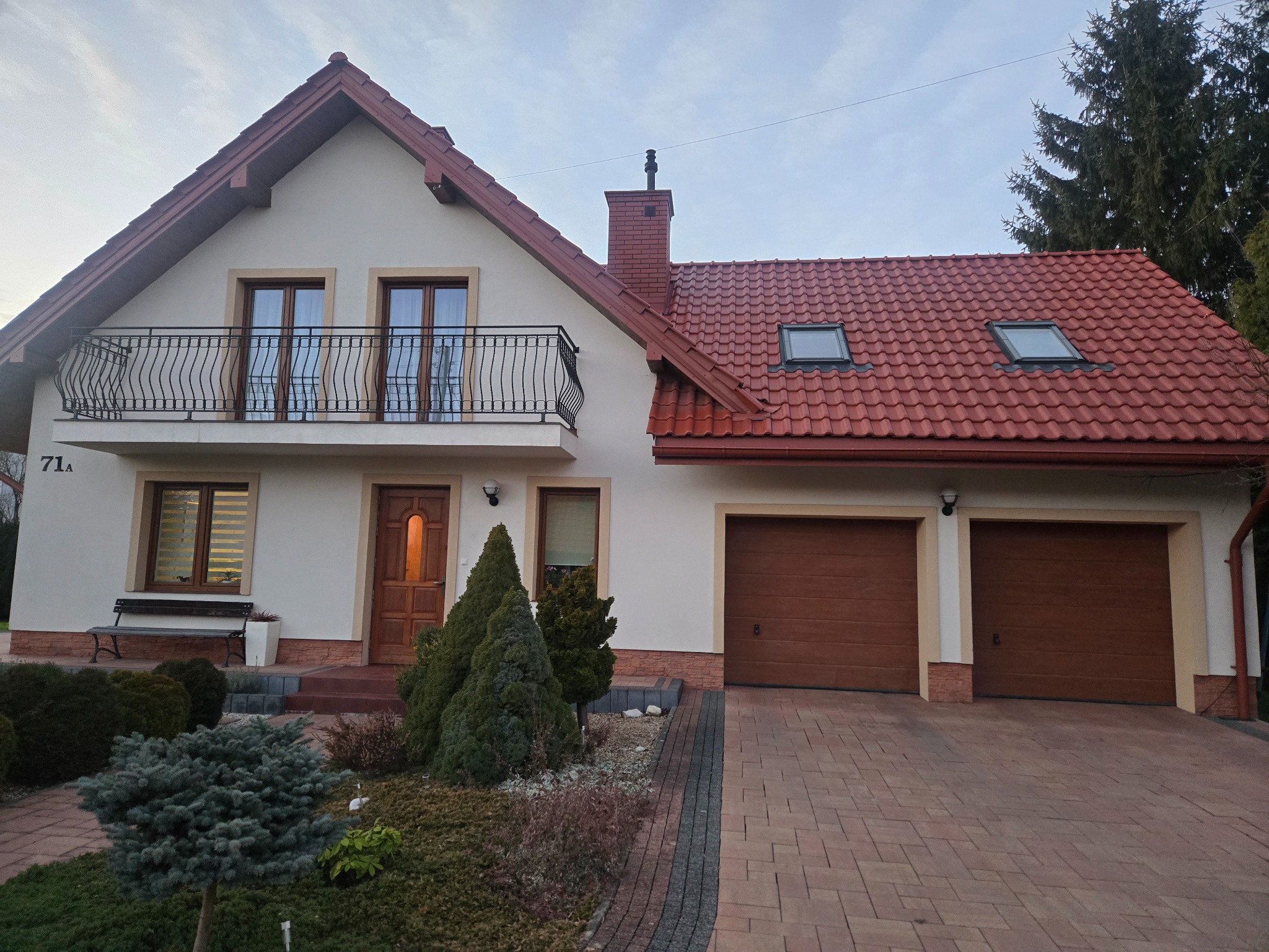 Dwukondygnacyjny dom z balkonem i dwoma garażami, wykończony czerwoną dachówką i jasną elewacją. Widoczne elementy architektury i zagospodarowania terenu.