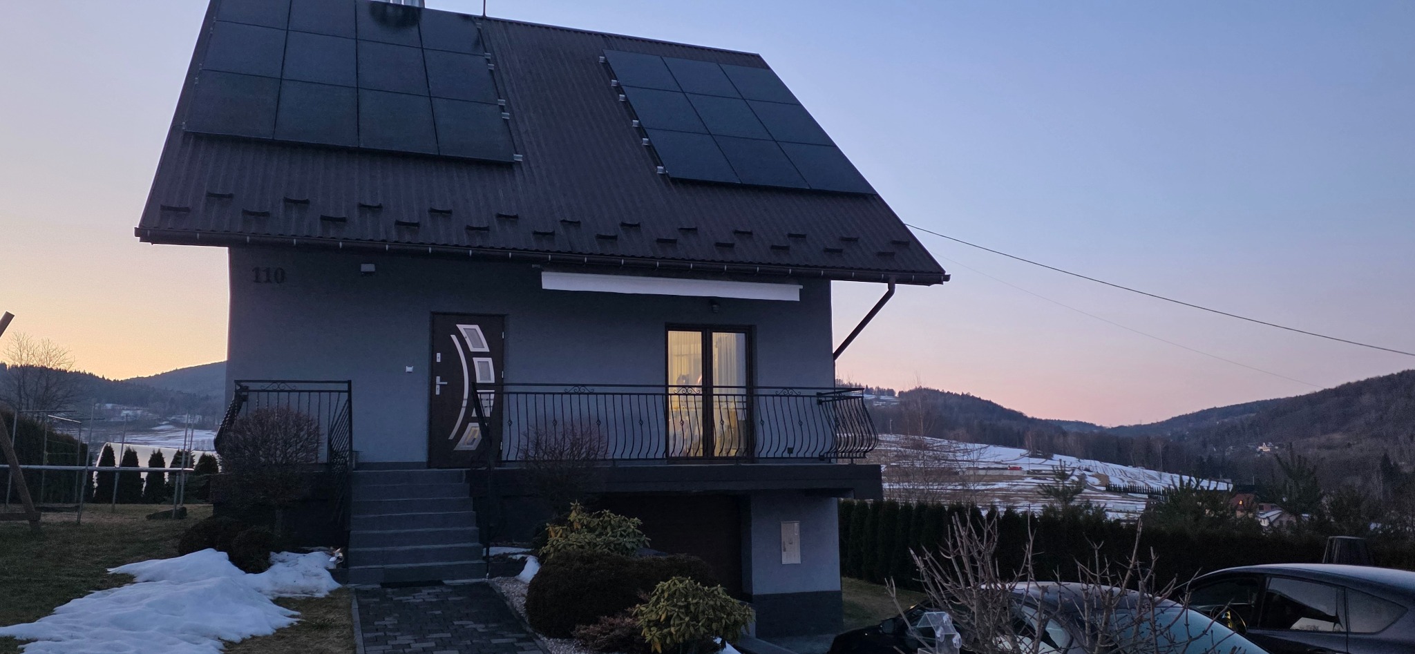Szary dom jednorodzinny z panelami słonecznymi na dachu, numer 110, balkon z kutą balustradą, w tle góry i zachodzące słońce.