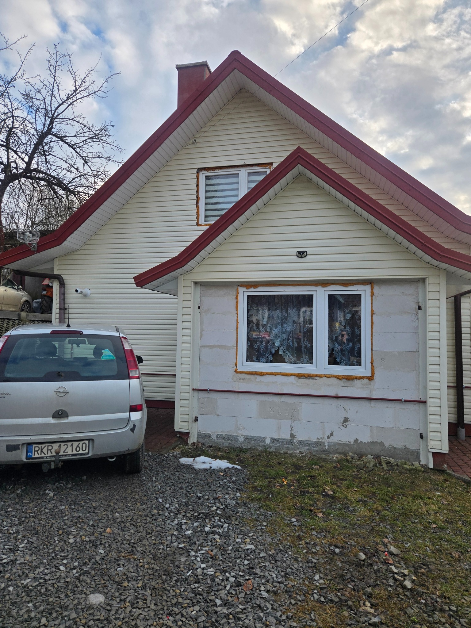 Dom z sidingiem i nowym oknem w trakcie montażu, widoczna piana montażowa. Na podjeździe stoi srebrny samochód osobowy. Pochmurne niebo w tle.