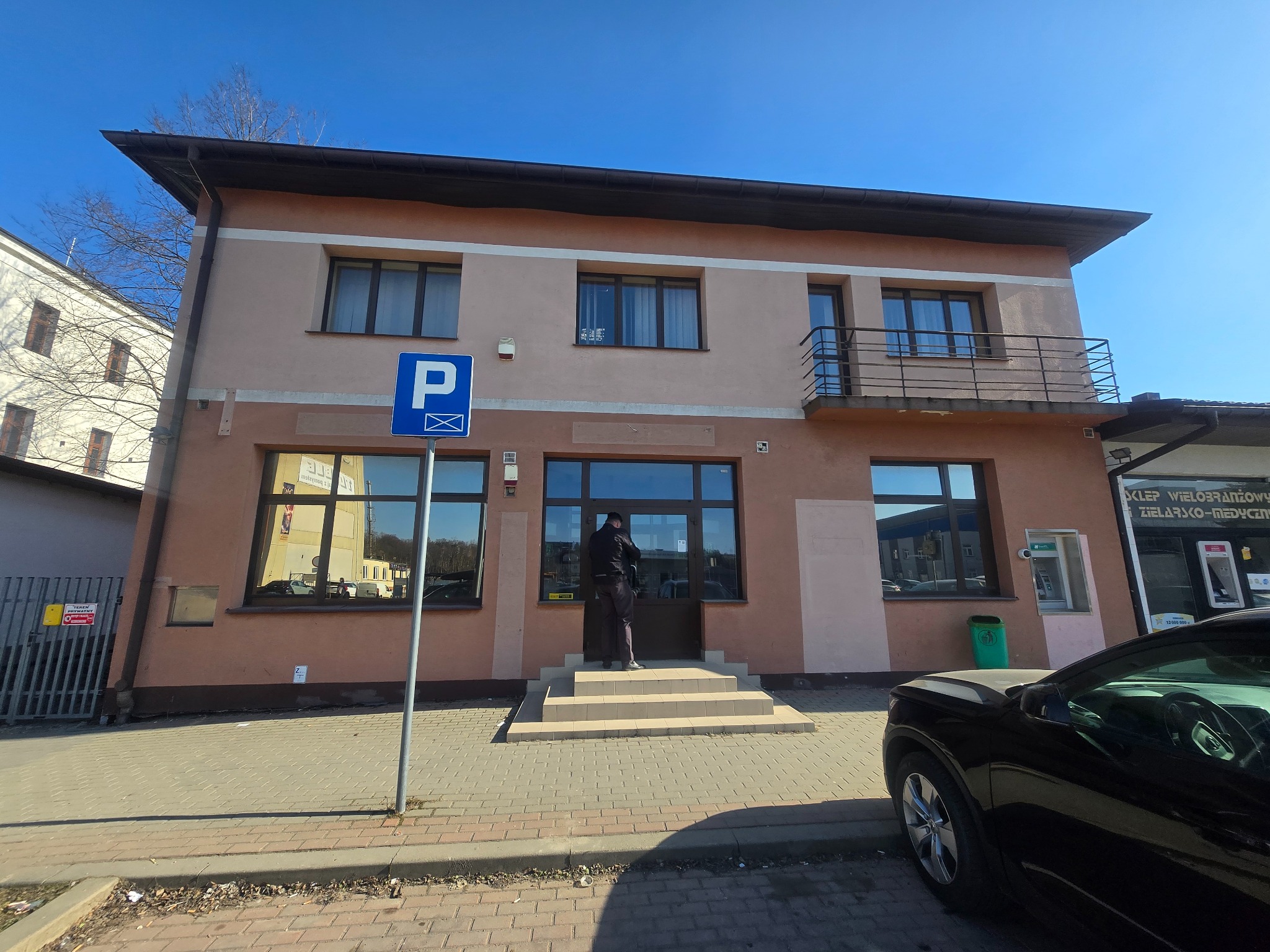 Dwupiętrowy budynek z balkonem i znakiem parkingu. Osoba stoi przed wejściem. Widoczny fragment chodnika i samochodu. Słoneczny dzień.
