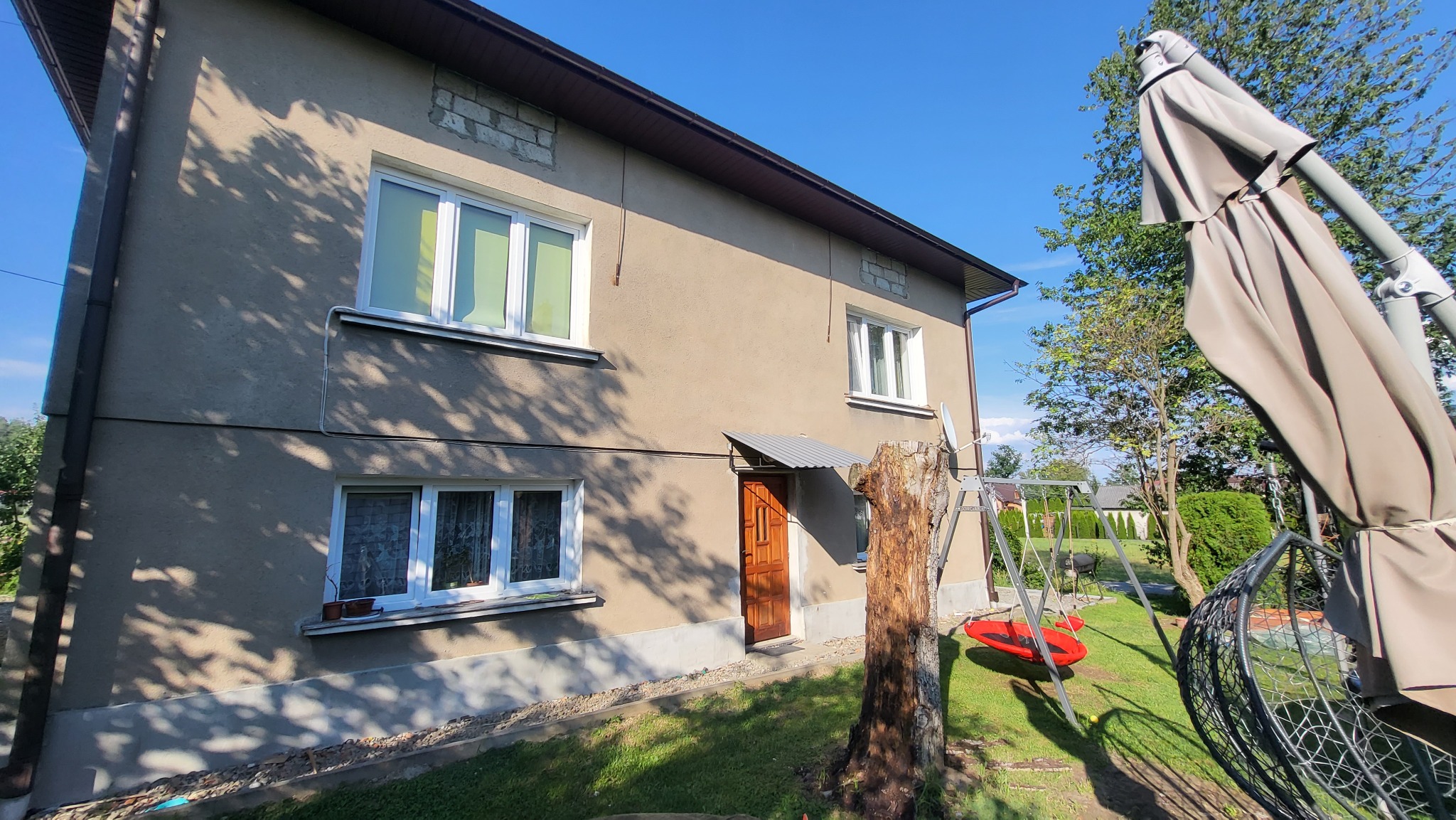 Dwukondygnacyjny dom z beżową elewacją, białymi oknami i brązowymi drzwiami wejściowymi. Widoczne drzewo, huśtawka i parasol ogrodowy. Błękitne niebo w tle.