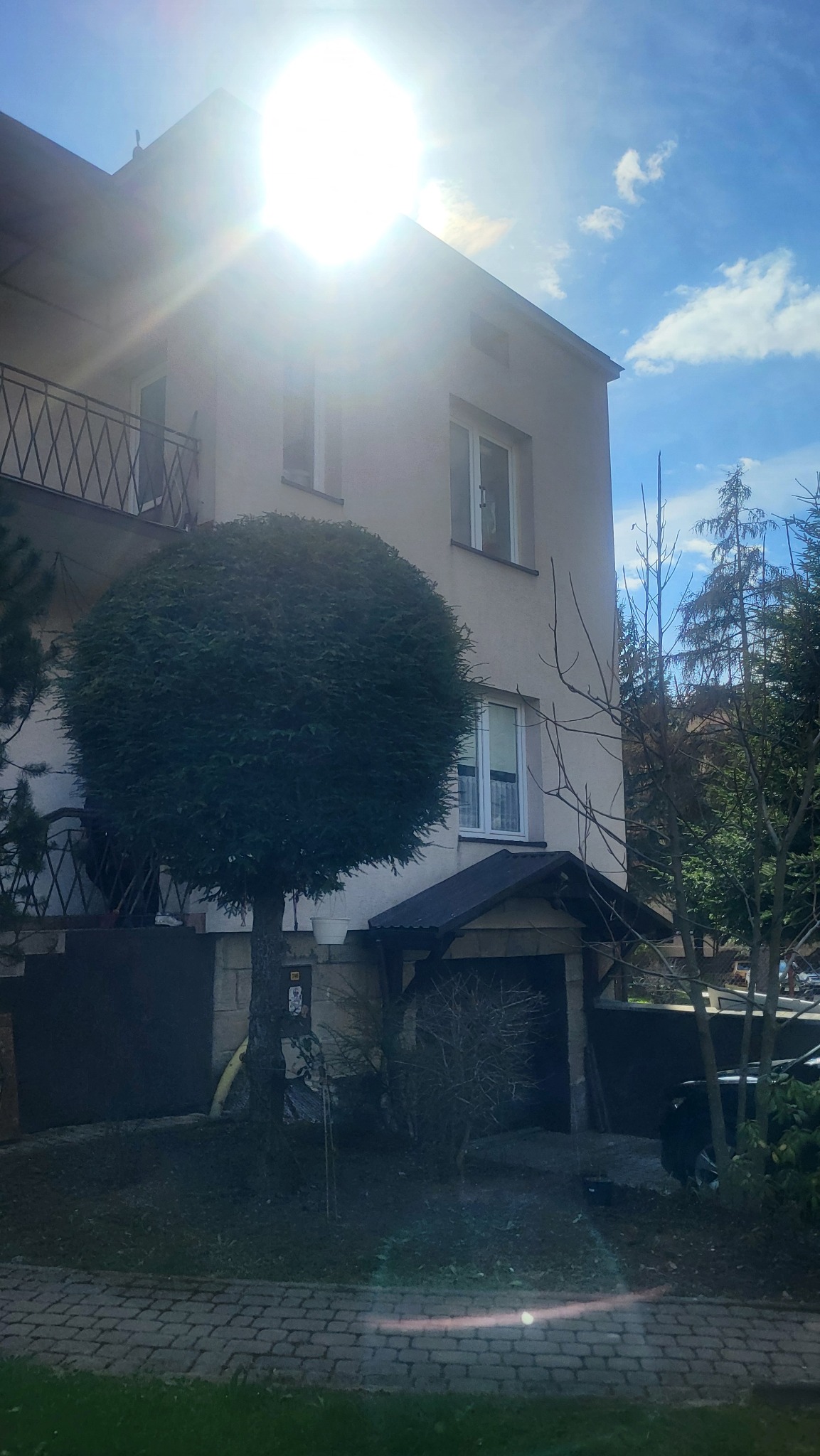 Dwupiętrowy dom z balkonem i kulistym drzewem na pierwszym planie, oświetlony silnym słońcem. Widoczne okna, brama wjazdowa i fragment nieba z chmurami.