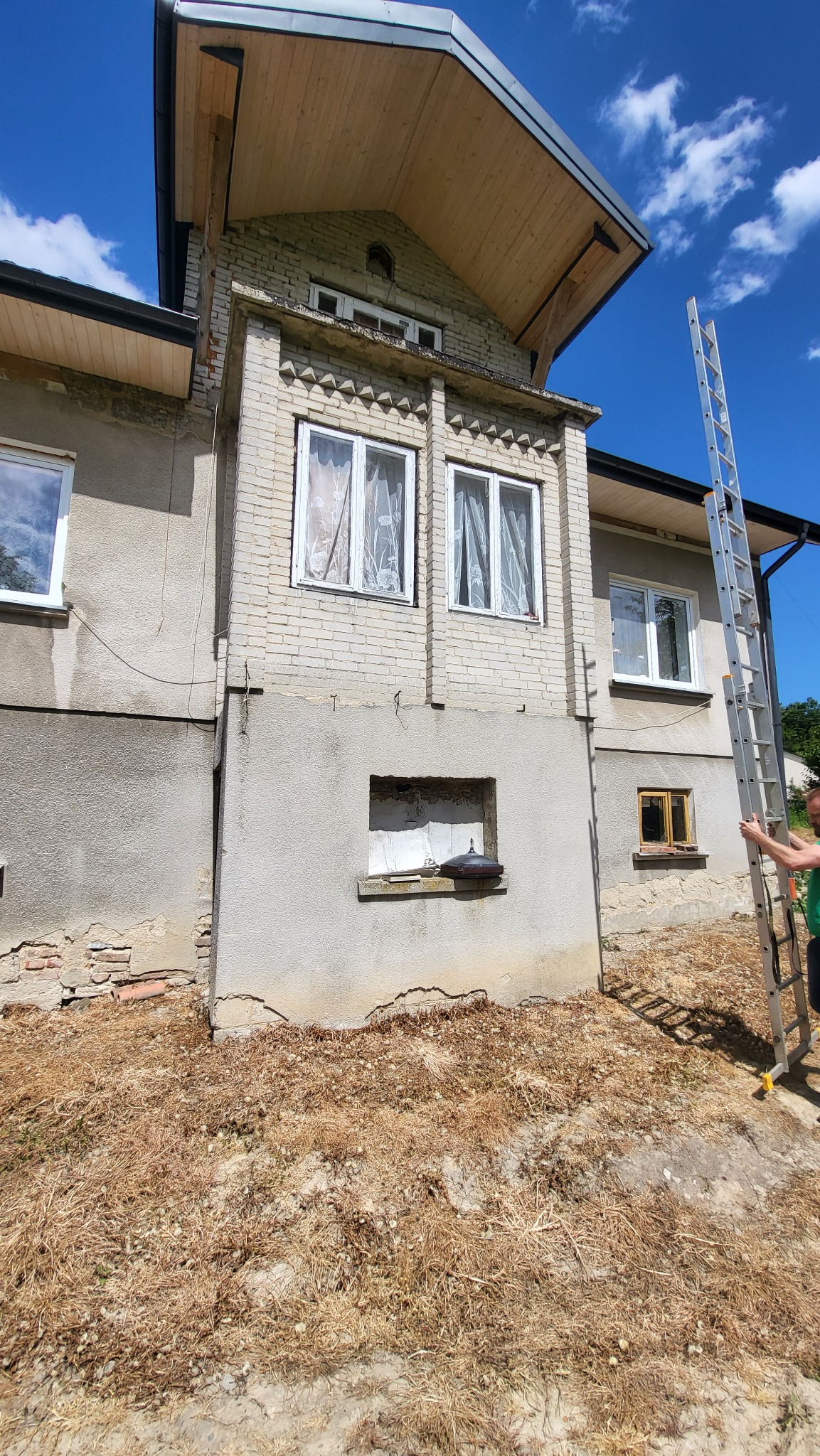 Dwukondygnacyjny dom z ceglaną elewacją i drewnianym dachem, z przystawioną drabiną, sugerujący prace remontowe lub inspekcję budynku.