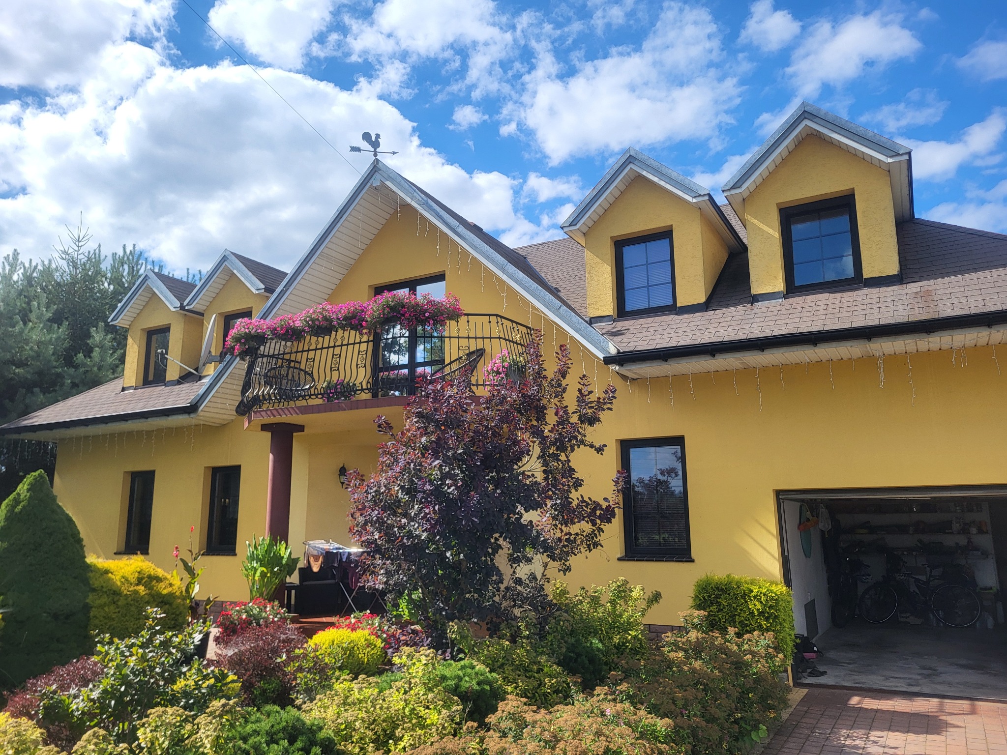 Żółty dom z balkonem ozdobionym kwiatami, widok z ogrodu. W tle błękitne niebo z chmurami. Otwarty garaż z rowerami.