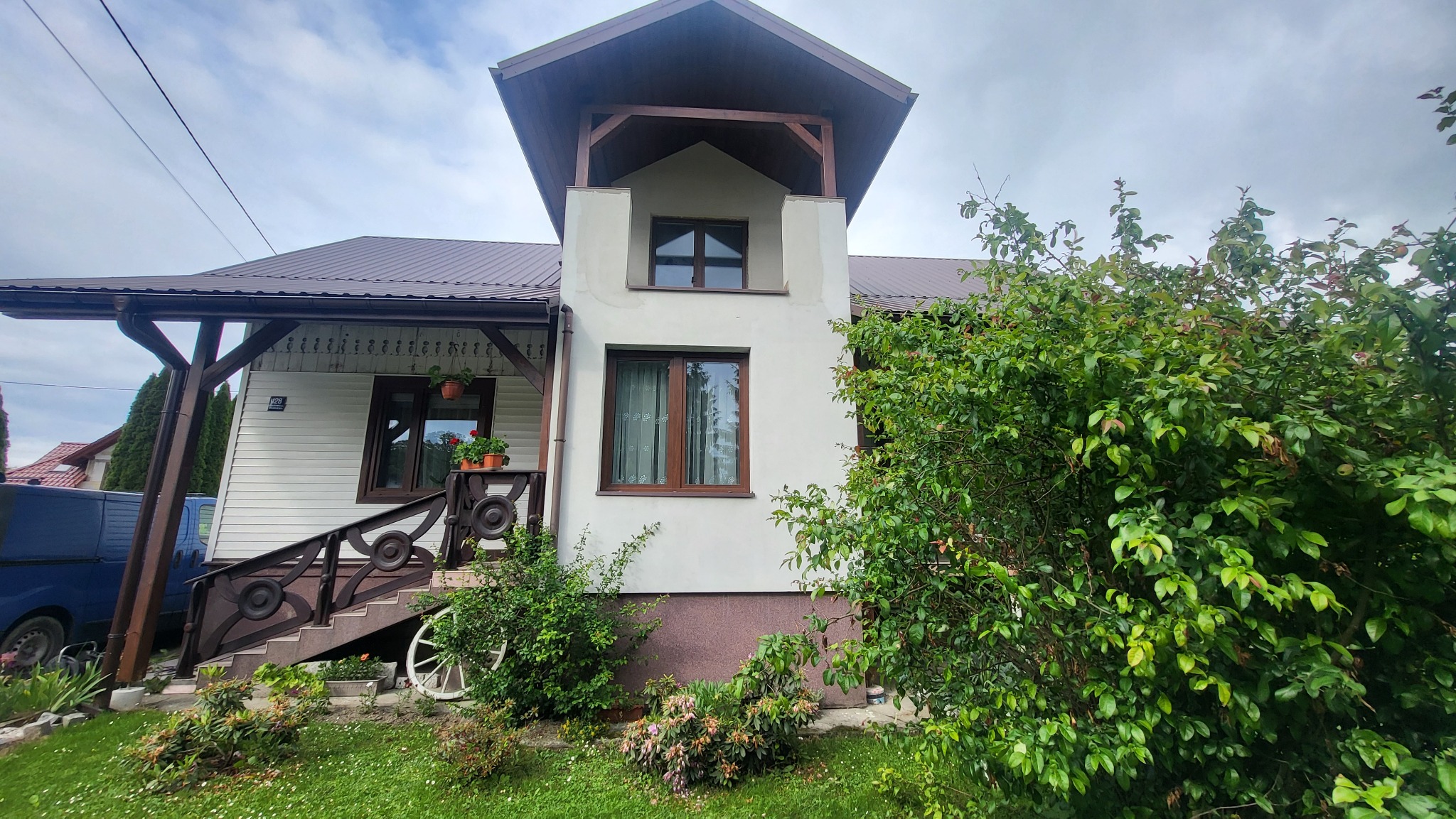 Dwukondygnacyjny dom z brązowym dachem i drewnianymi elementami, widoczny z poziomu trawnika, z bujną zielenią po prawej stronie.