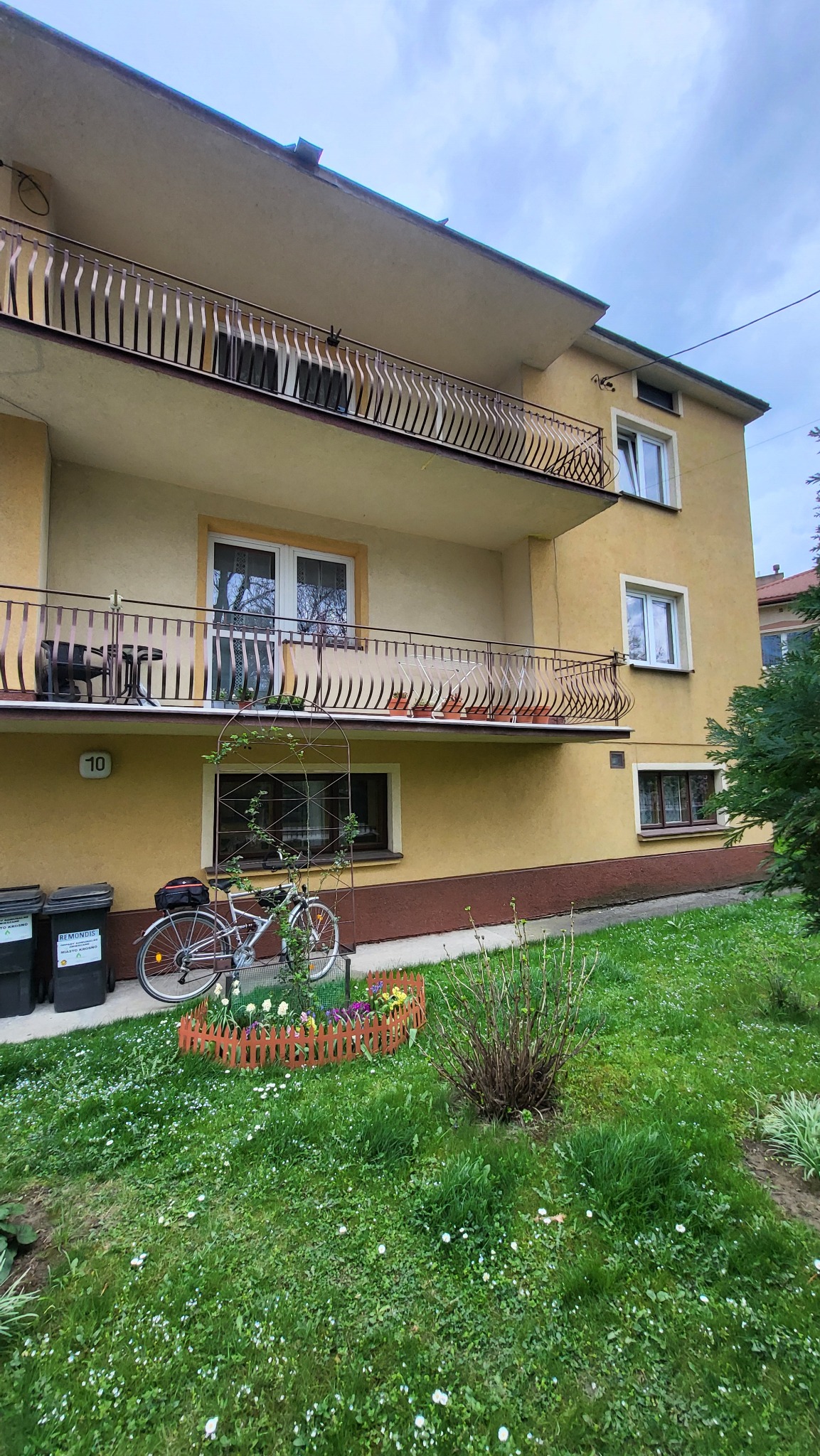 Żółty budynek z balkonami i numerem 10, rower oparty o ścianę, mały ogródek z kwiatami i zielony trawnik z białymi kwiatami. Widok z dołu na budynek.