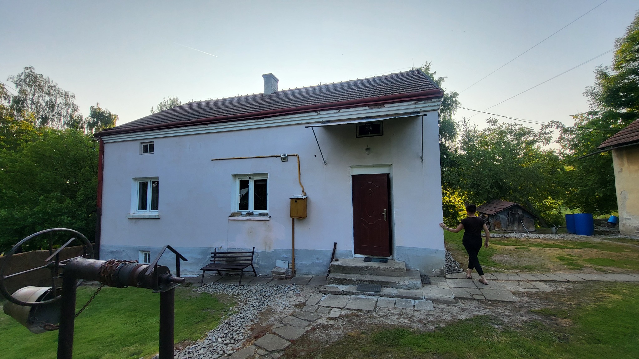 Starszy, jednopiętrowy dom z brązowymi drzwiami wejściowymi i studnią na pierwszym planie. Kobieta w oddali. Otoczenie: zieleń, drzewa.