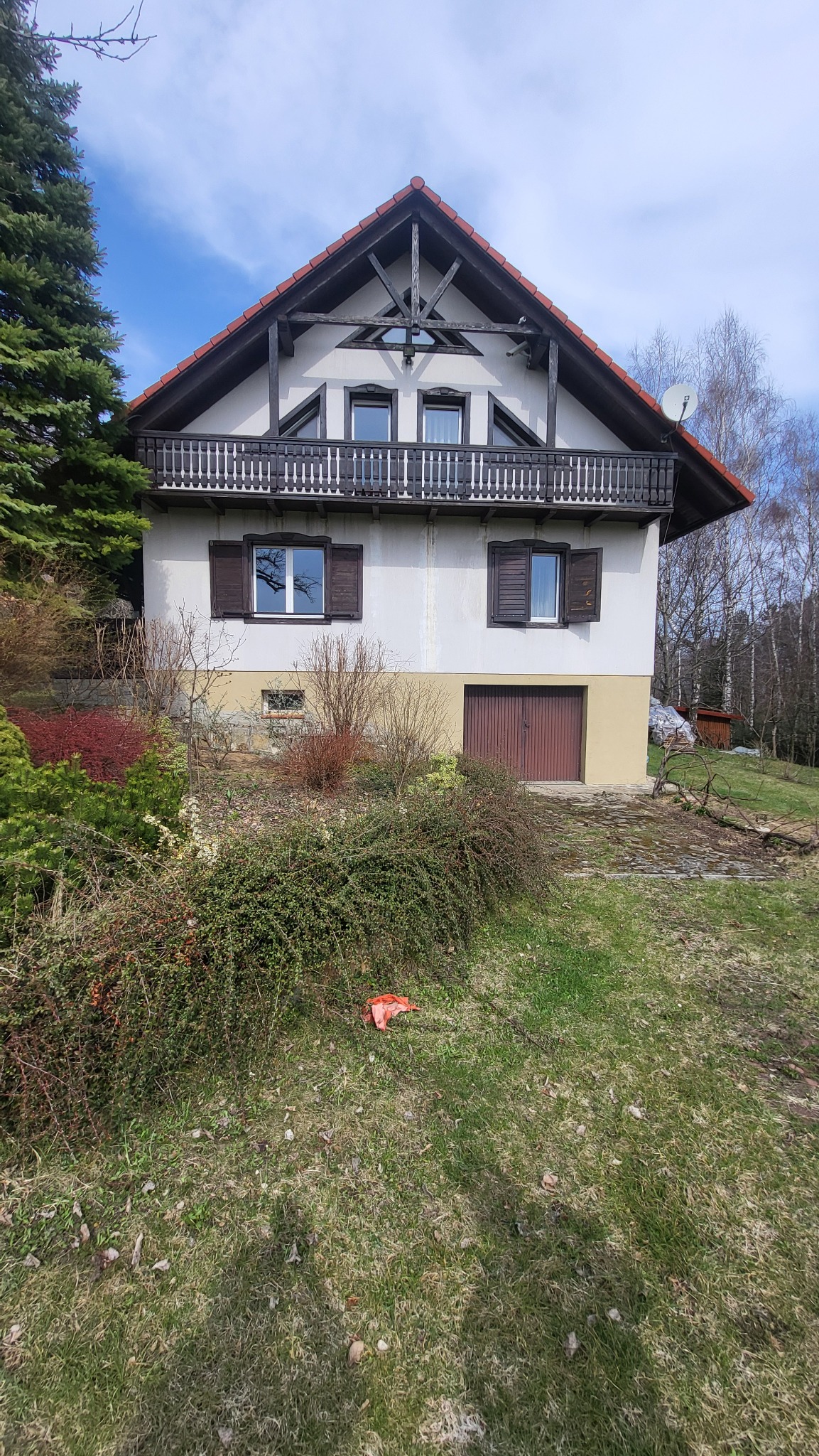 Dwukondygnacyjny dom z balkonem i brązowymi okiennicami, otoczony zielenią. Widoczna antena satelitarna i garaż. Ujęcie z poziomu gruntu.