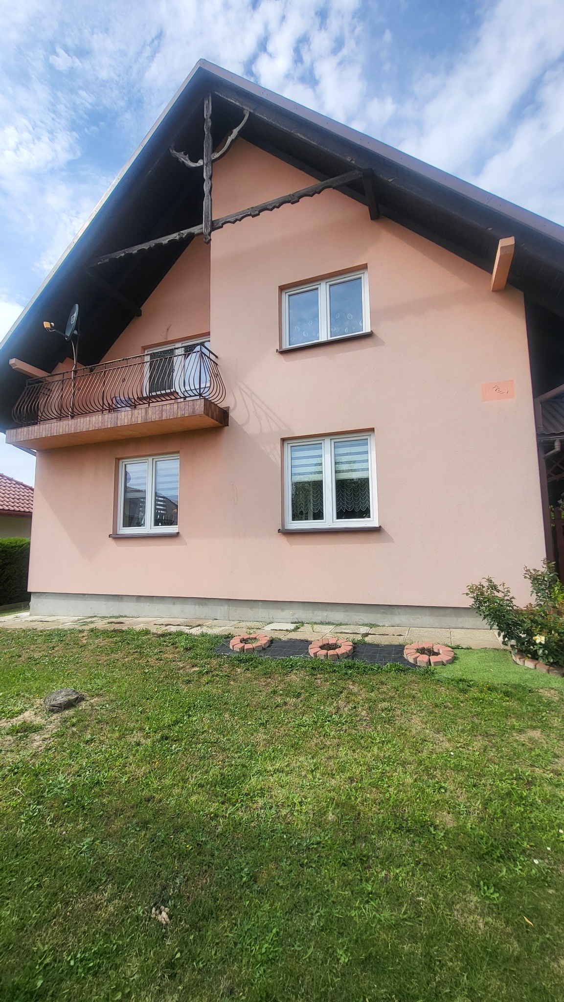 Dwukondygnacyjny dom z balkonem i beżową elewacją, widoczny z poziomu trawnika. Charakterystyczne drewniane zdobienia pod dachem. Okna z białymi ramami.